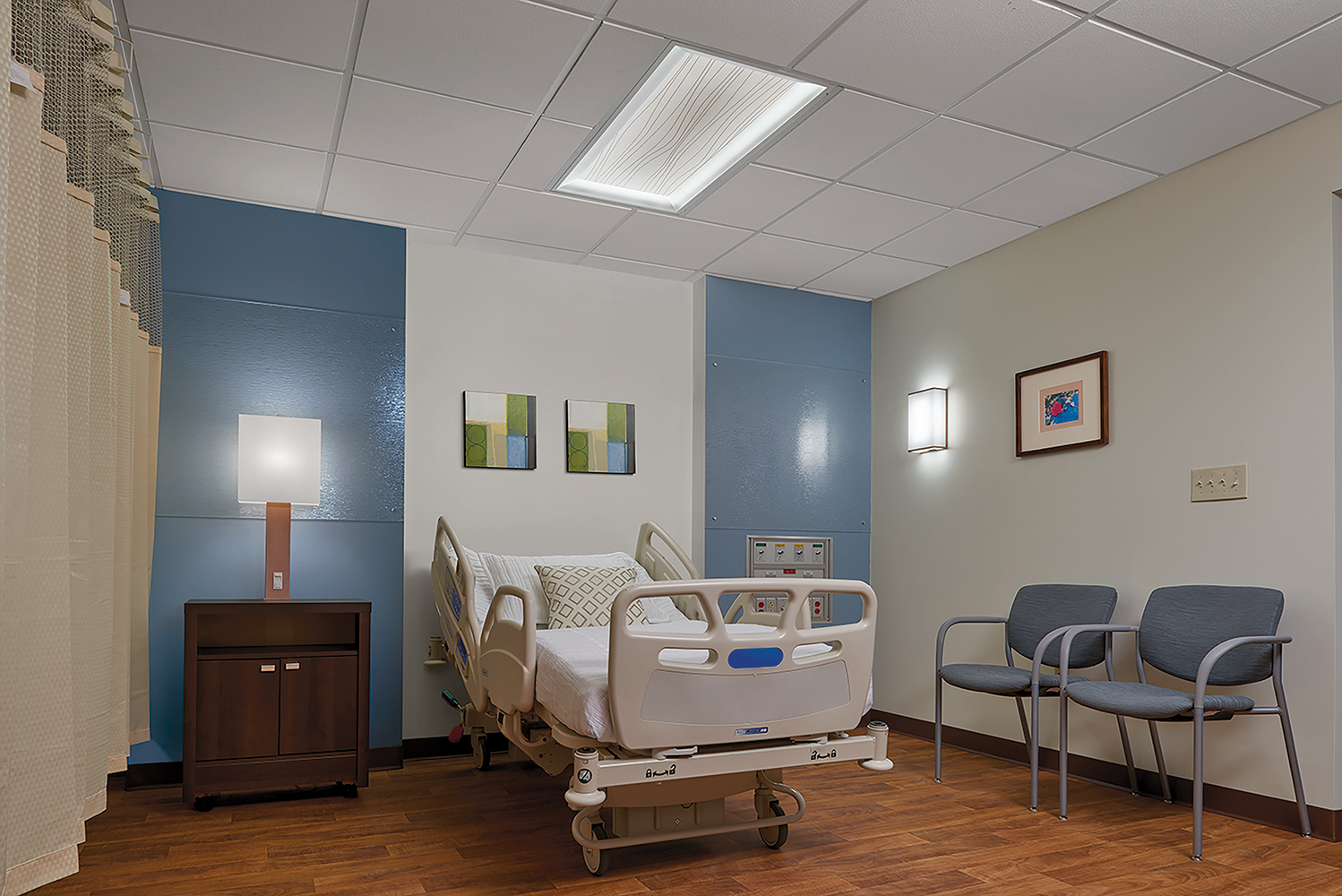 Serenity patient room lighting fixtures illuminate a hospital bed from a table lamp, overbed luminaire, and wall sconce.