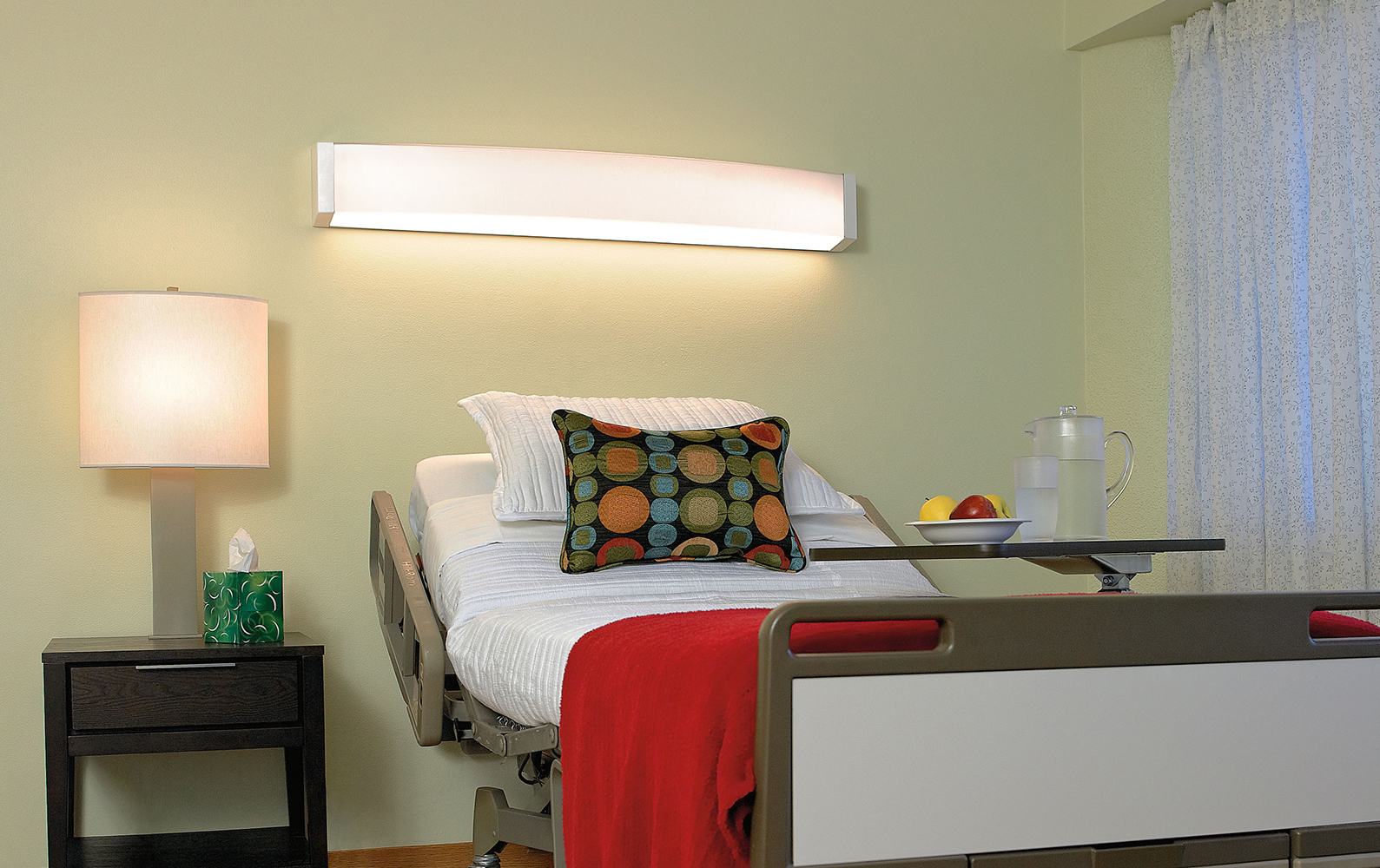 Unity medical lighting fixtures in headwall and table lamp versions, over a patient bed with a side table and colorful pillow.