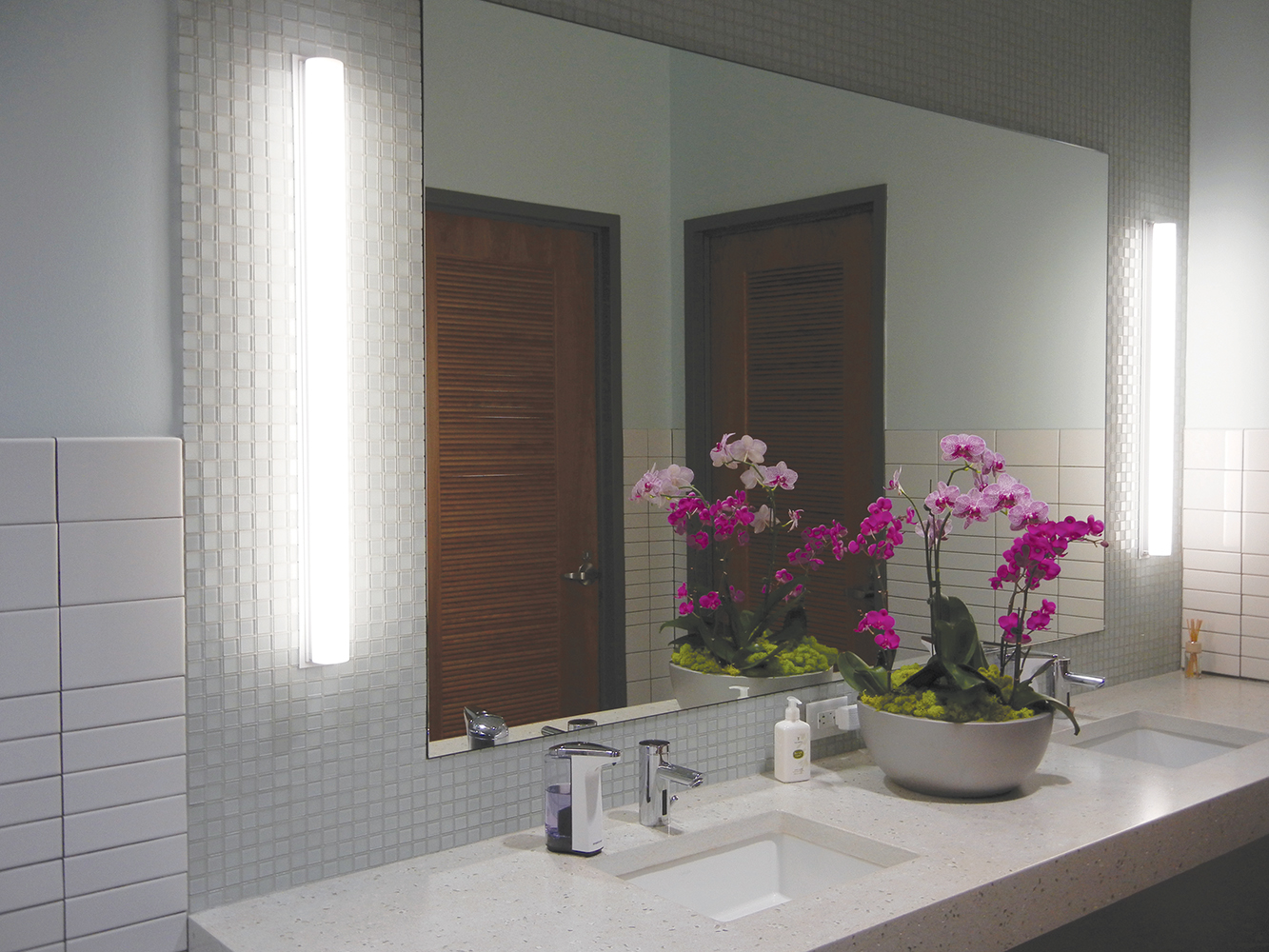 Voila modern vanity light fixtures illuminate mirrors in sleek, clean restroom with a bowl of purple flowers between two sinks.