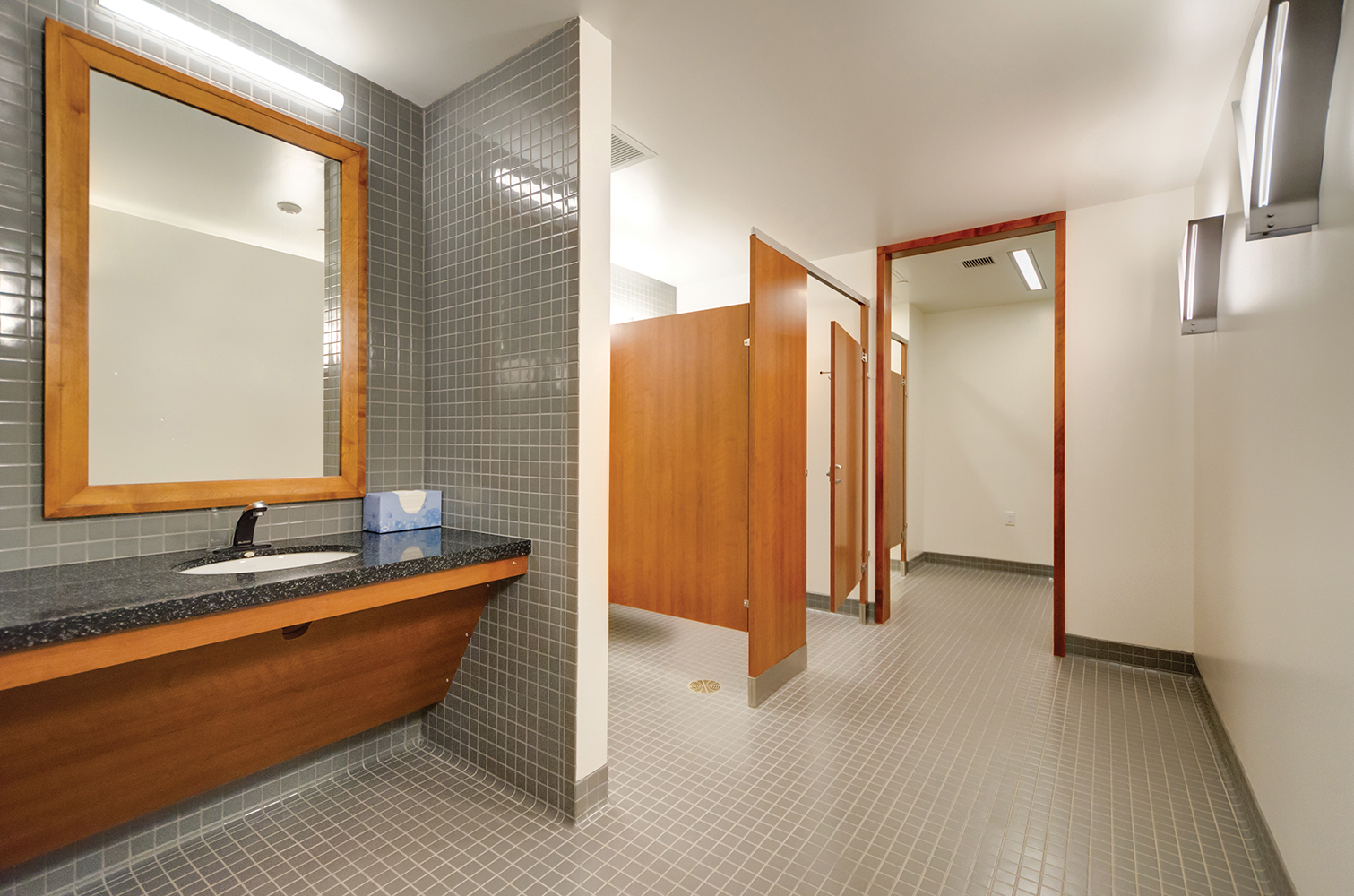 Voila modern vanity light fixtures illuminate a public restroom, mounted horizontally above a wood-framed mirror.